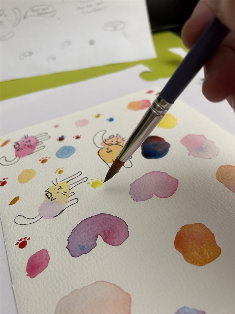 A hand holding a paintbrush over a sheet of watercolor paper with colorful paint swatches and small ink-drawn cats.
