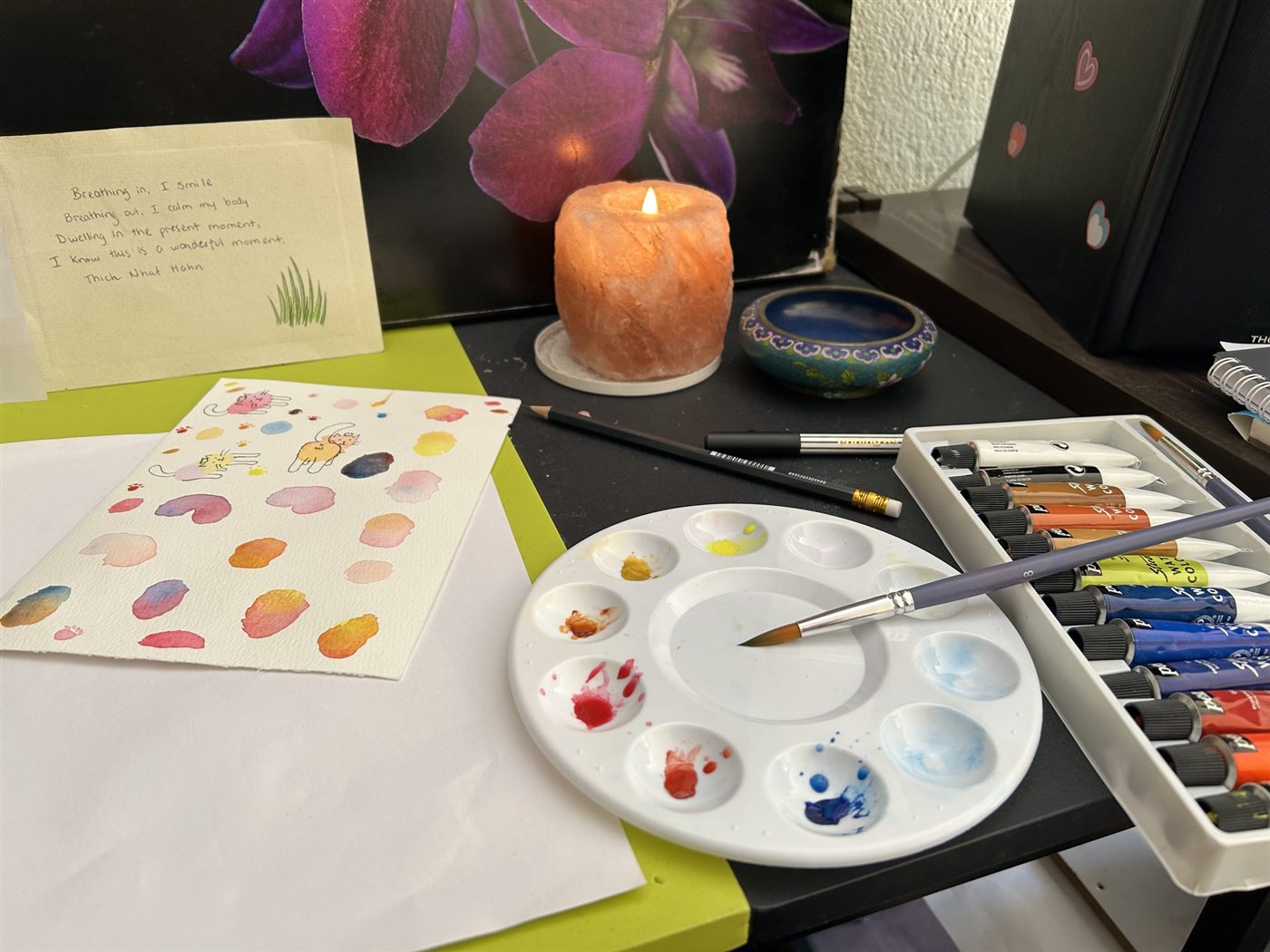 A painting table with a lit salt-crystal candle, a paint palette, tubes of watercolor, and a painted sheet of cats and swatches.